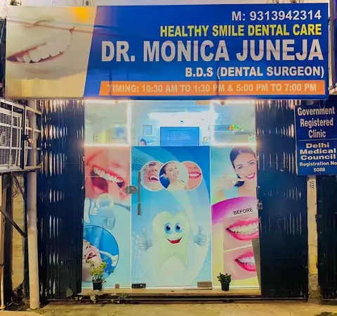 The clinic's glass entrance at night, lit by a blue signboard reading Healthy Smile Dental Care, Dr. Monica Juneja, B.D.S. (Dental Surgeon), with a Delhi Medical Council registration plate beside the door.