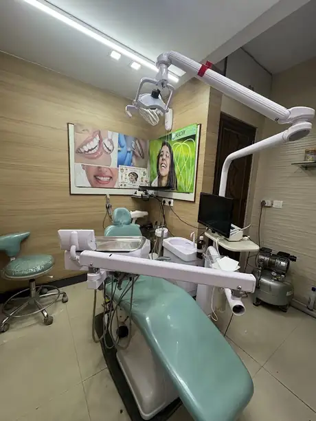 A treatment room with a mint-green dental chair, an overhead operating light, a wall-mounted monitor and dental charts on the wood-panelled wall.