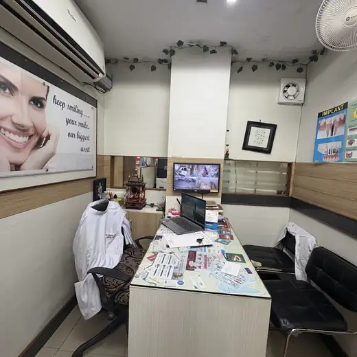 A consulting desk with a laptop and printed leaflets, a white coat over the chair behind it and black waiting chairs opposite.